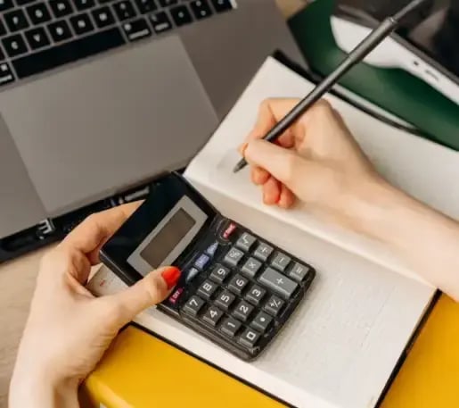 Person calculating figures with a black calculator and recording data in a notepad, with a laptop visible in the background, illustrating accounting or financial tasks.