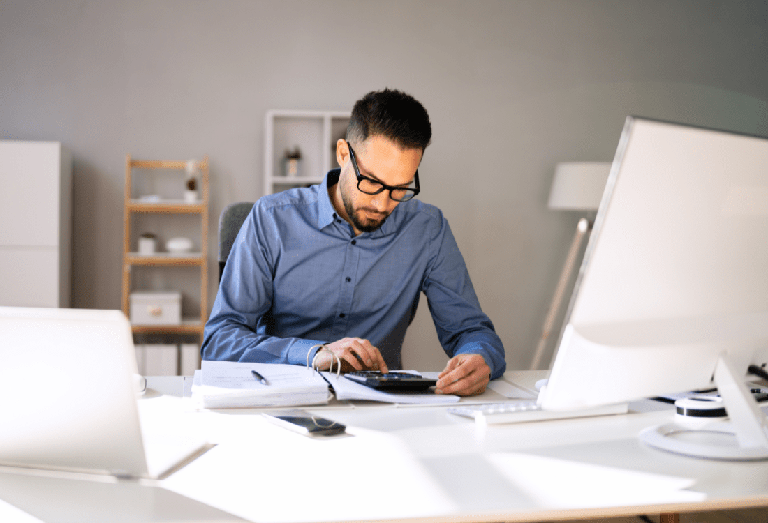 An outsourced bookkeeper working at his desk with a desktop, laptop, ring binder, and using a calculator who is gathering accurate and reliable information for a small business to save them time and money. 