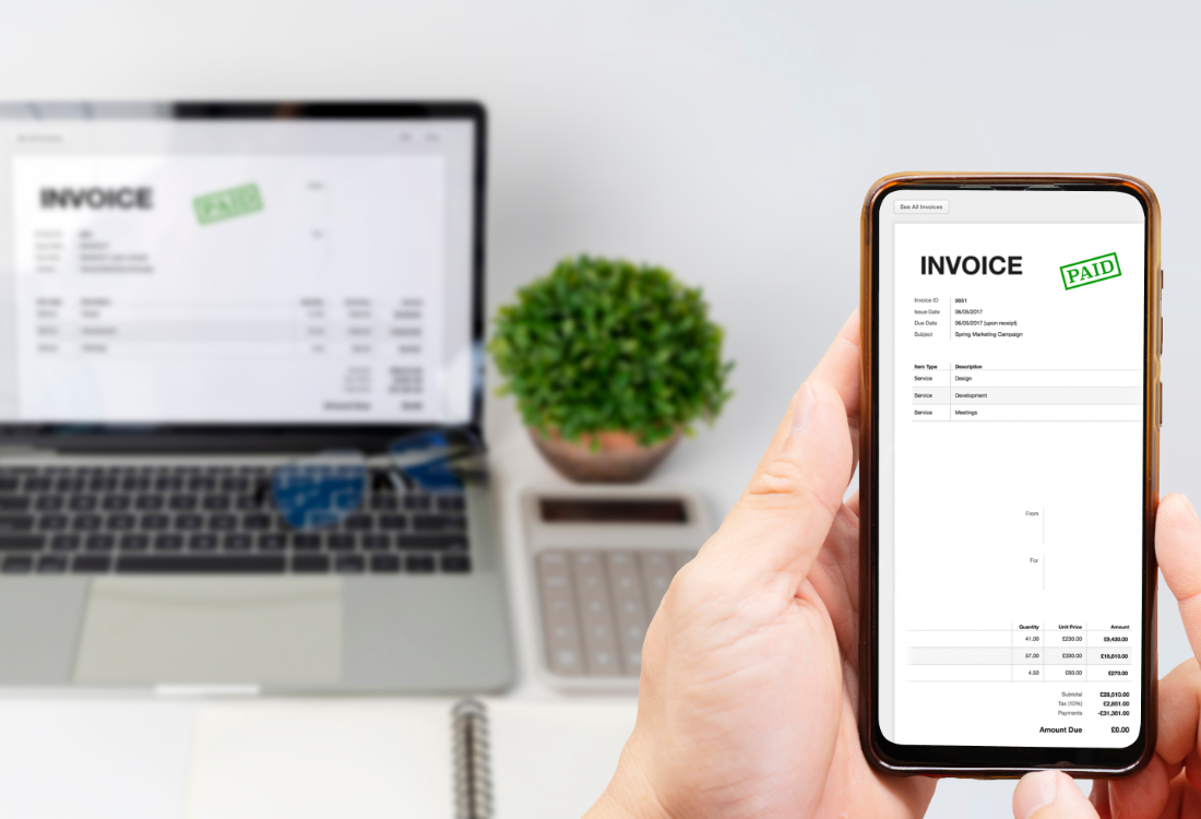 A person holding a smartphone displaying a paid digital invoice, with the same invoice open on a laptop in the background, representing Making Tax Digital and online accounting. 
