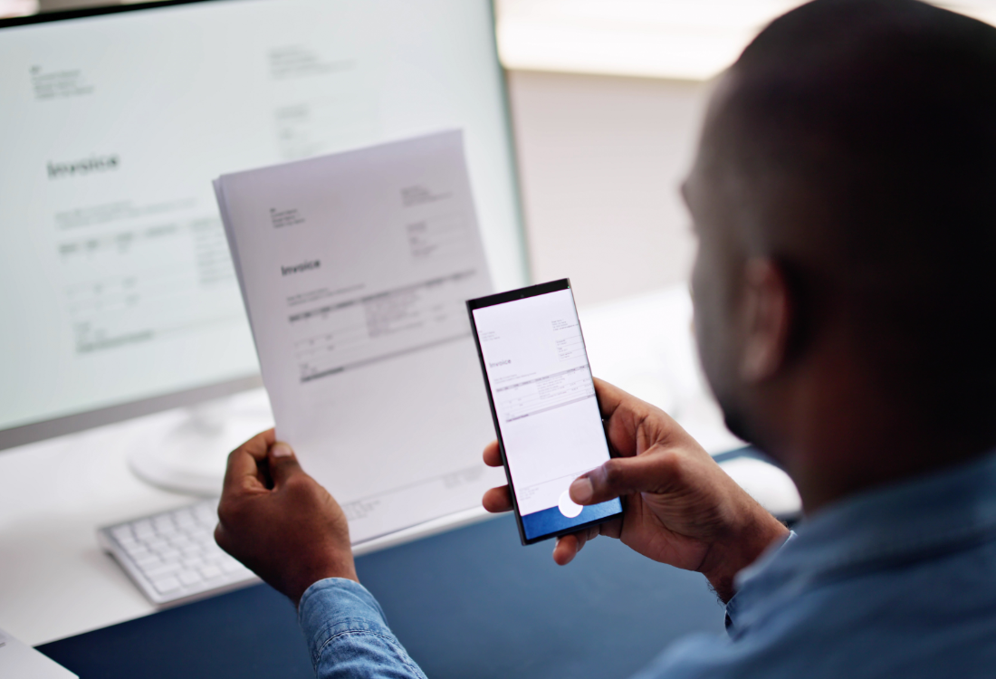 A person scanning a paper invoice with a smartphone while working on a computer, illustrating digital record-keeping for Making Tax Digital compliance. 