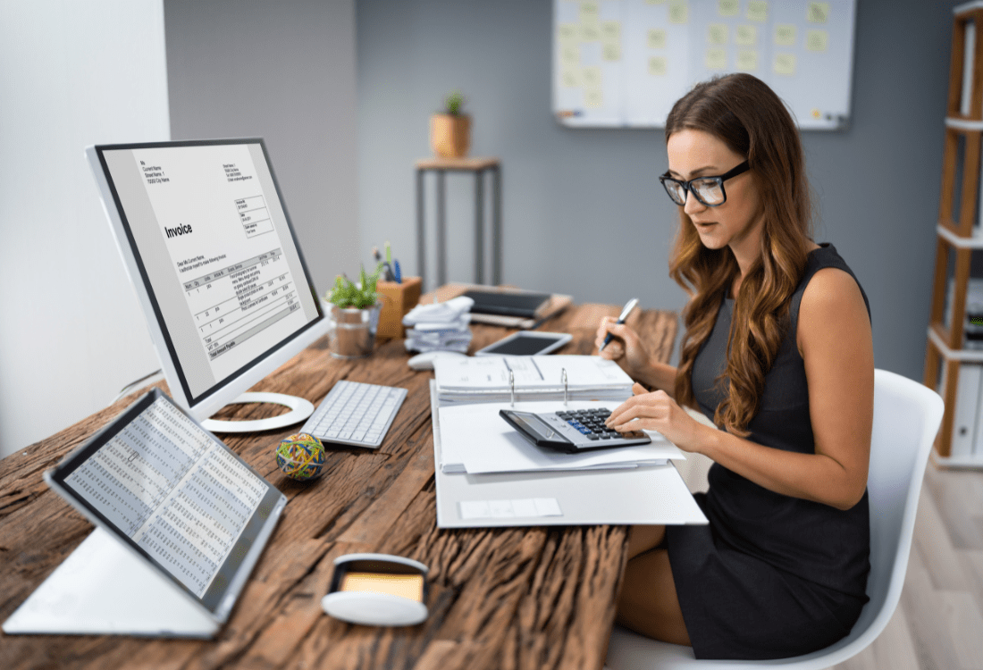 An accountant sitting at her desk with invoices on her desktop, a spreadsheet on her tablet, calculating the tax using a full ring binder and calculator for a business. 